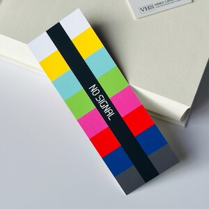 May include: A colorful bookmark with a retro TV test pattern design. The bookmark features a black vertical stripe with the words "NO SIGNAL" in white. The background is a mix of bright colors including yellow, blue, green, pink, and red.