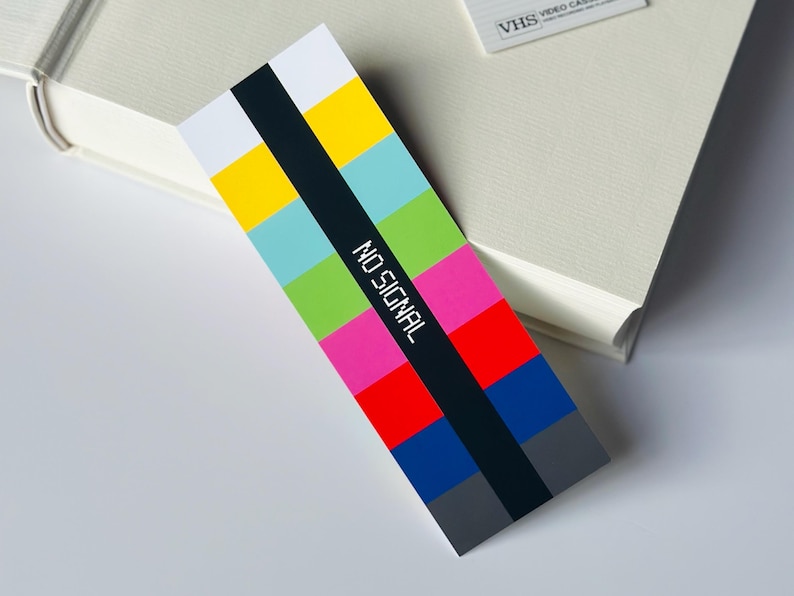 May include: A colorful bookmark with a retro TV test pattern design. The bookmark features a black vertical stripe with the words "NO SIGNAL" in white. The background is a mix of bright colors including yellow, blue, green, pink, and red.