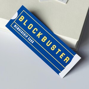 May include: A blue Blockbuster membership card with yellow text. The card reads "BLOCKBUSTER" and "MEMBERSHIP CARD". The card is rectangular with a white border and is placed on a white surface.