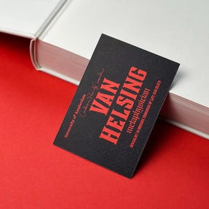K&ouml;nnte beinhalten: Eine schwarze Visitenkarte mit dem Namen "Van Helsing" in roter, fetter Schrift. Die Karte enth&auml;lt auch den Titel "Metaphysiker" und den Text "Spezialist f&uuml;r obskure Bereiche von Leben und Tod". Die Karte liegt auf einer roten Oberfl&auml;che.