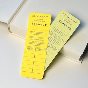 May include: Two yellow library cards with black text, one labelled "Spencer" and the other "Zachary." The cards have a vintage design with space for book titles and due dates. They rest on a white book.