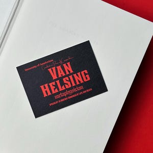 K&ouml;nnte beinhalten: Eine schwarze Visitenkarte mit dem Namen "VAN HELSING" in Rot, vor einem wei&szlig;en Hintergrund. Die Karte enth&auml;lt auch den Text "Metaphysiker" und "Spezialist f&uuml;r obskure Ecken des Lebens und des Todes."