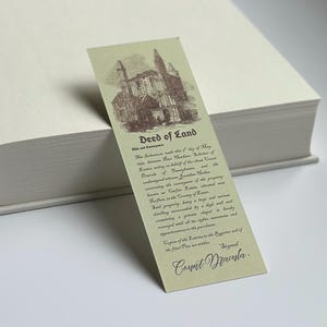 May include: A vintage-style bookmark with a sepia-toned illustration of a gothic building. The bookmark features the text "Deed of Land" and detailed text below, signed "Count Dracula". The bookmark is resting on a thick, off-white book.