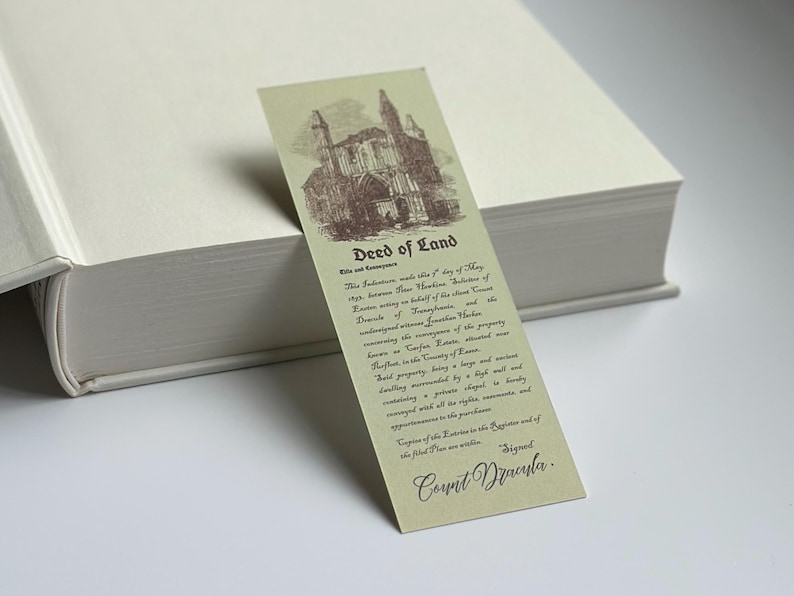 May include: A vintage-style bookmark with a sepia-toned illustration of a gothic building. The bookmark features the text "Deed of Land" and detailed text below, signed "Count Dracula". The bookmark is resting on a thick, off-white book.