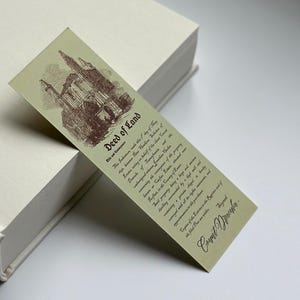 May include: A vintage-style bookmark with the text "Deed of Land" and a sepia-toned illustration of a building. The bookmark is light green and features detailed text attributed to "Count Dracula". It rests on a white book.