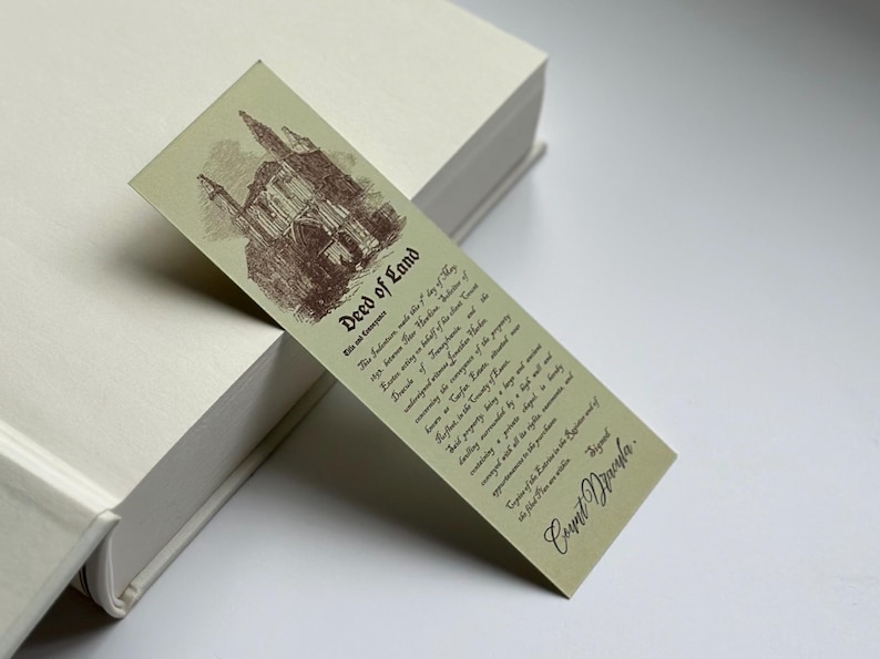 May include: A vintage-style bookmark with the text "Deed of Land" and a sepia-toned illustration of a building. The bookmark is light green and features detailed text attributed to "Count Dracula". It rests on a white book.