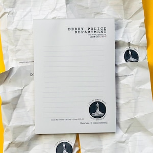 May include: A white notepad with horizontal lines, featuring the text "DERRY POLICE DEPARTMENT" and a case number. The notepad has a circular seal with a lighthouse image and the text "CITY OF DERRY, MAINE". The notepad is on a crumpled paper background.