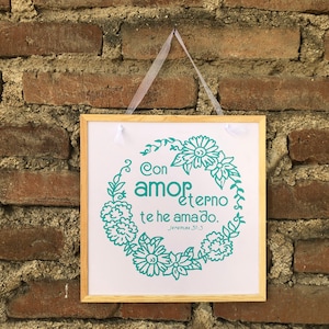 May include: A framed print with a white background and a teal green floral wreath. The wreath surrounds the text "Con amor eterno te he amado. Jeremiah 31:3".