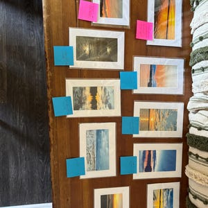 Op de afbeelding: Een verzameling ingelijste landschapskunstafdrukken met zonsondergang- en zeelandschapsscènes. De afdrukken tonen levendige kleuren van oranje, geel, blauw en grijs. De kunst wordt gepresenteerd op een houten oppervlak, met plaknotities bevestigd aan sommige frames.