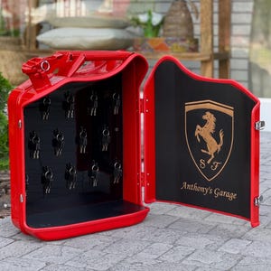 May include: A red metal key holder shaped like a gas can, with a black interior and a door featuring a gold emblem and the text "Anthony's Garage". The key holder is open, revealing multiple keys.