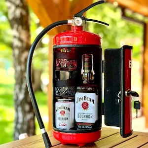 May include: A red fire extinguisher converted into a drinks cabinet. The open door reveals a bottle of Jim Beam Bourbon, a can of Jim Beam Bourbon & Cola, and two crystal glasses. The interior of the cabinet is black.