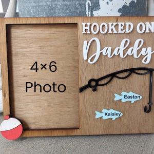 May include: Wooden photo frame with a 4x6 photo space. The frame is decorated with the words "Hooked on Daddy", a fishing rod, and two blue fish with the names "Easton" and "Kaisley". A red and white bobber is in the lower left corner.