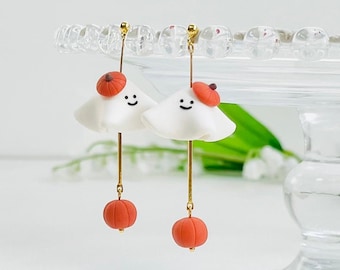 May include: A pair of whimsical Halloween earrings. Each earring features a white ghost with a smiling face, a small orange beret, and a dangling orange pumpkin. The earrings are suspended from gold-colored posts.