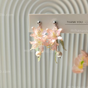 May include: A pair of drop earrings featuring translucent pale pink lily flowers with delicate yellow stamens, green leaves, and a single white pearl bead hanging from each silver-toned post. The earrings are attached to a white card with the text THANK YOU.