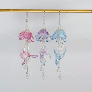 Puede incluir: Tres pares de pendientes colgantes con una parte superior en forma de flor y una larga cinta translúcida retorcida que cuelga debajo. Los pendientes están disponibles en rosa, morado y azul.