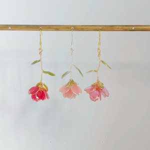 Könnte beinhalten: Drei hängende Blumenohrringe mit durchscheinenden rosa und roten Blütenblättern sowie grünen Blättern an dünnen Drahtstielen. Die linken und rechten Ohrringe haben goldfarbene Haken, während der mittlere Ohrring einen silberfarbenen Haken besitzt.