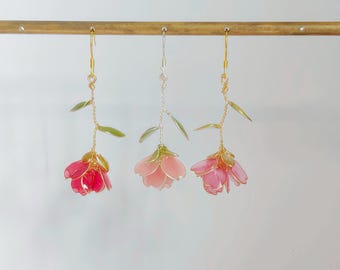 May include: Three dangling flower earrings featuring translucent pink and red petals with green leaves on thin wire stems. The left and right earrings have gold-toned hooks, while the centre earring has a silver-toned hook, all hanging from a horizontal bar.