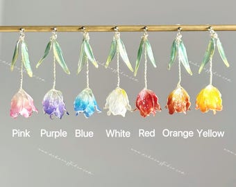 May include: Seven tulip-shaped earrings hanging from a gold-toned bar. Each earring features a translucent flower in pink, purple, blue, white, red, orange, or yellow, with two green leaves and a silver-toned stem. Text labels identify each colour.