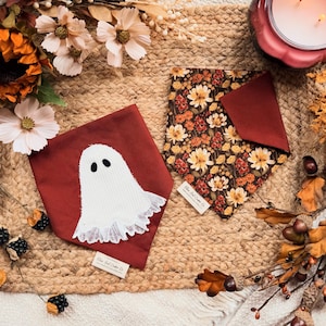 May include: Two triangular pet bandanas. One is solid dark red with a white corduroy ghost applique featuring a lace hem. The other is a dark brown fabric with a repeating pattern of orange, yellow, and white autumn flowers and green leaves.