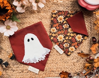 May include: Two triangular pet bandanas. One is solid dark red with a white corduroy ghost applique featuring a lace hem. The other is a dark brown fabric with a repeating pattern of orange, yellow, and white autumn flowers and green leaves.