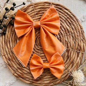 May include: Two orange fabric hair bows with a pearl trim. The larger bow is tied in a loop and the smaller bow is tied in a knot. The bows are displayed on a woven basket.
