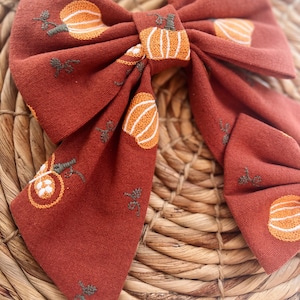 May include: A large, rust-colored fabric bow with a pumpkin pattern. The bow is made of a soft, textured fabric and features embroidered pumpkins in shades of orange and white.