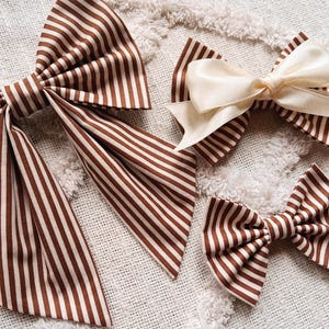 May include: Several fabric bows in varying sizes. The bows feature a brown and white striped pattern. One bow is solid cream-colored. The bows are arranged on a textured, off-white surface. The bows are likely hair accessories.