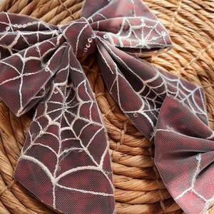 May include: A large red and white bow with a spiderweb design and silver sequins. The bow is made of tulle and is perfect for adding a touch of Halloween spirit to any outfit.