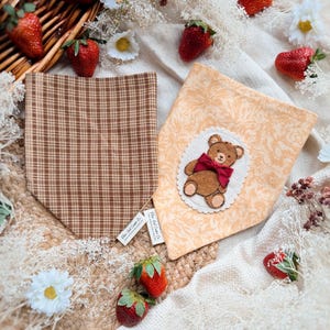 May include: Two pet bandanas. One is brown plaid, the other is peach with a teddy bear applique. Strawberries and daisies surround the bandanas. The bandanas are on a white cloth.