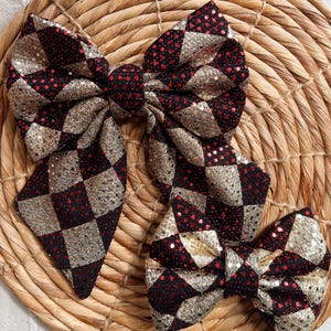 May include: Two red and gold checkered fabric hair bows. The larger bow is tied in a large loop with two long tails. The smaller bow is tied in a smaller loop with two shorter tails.