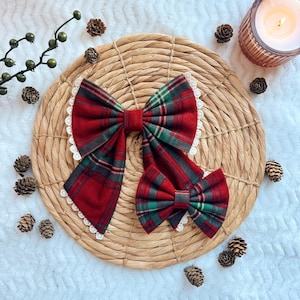May include: Two red plaid bows with green and black accents, one large and one small, are displayed on a woven placemat. The bows have white lace trim. Pinecones and a lit candle in a glass holder are in the background.