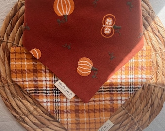 May include: Two dog bandanas, one with a red background and embroidered pumpkins, and the other with a plaid pattern in orange, white, and black. Both bandanas have a small label that reads "Blue Owl Crafts Co."
