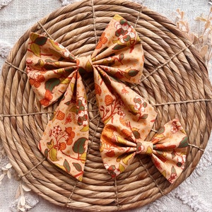 May include: Three fabric hair bows with a floral pattern in shades of orange, brown, and green. The bows are arranged on a woven straw surface.