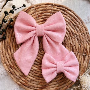 May include: Two pink sequined hair bows, one large and one small, on a woven straw background.