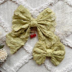 May include: A large, light green fabric bow with ruffled edges and a smaller bow attached. A gold ring with two red cherries hangs from the center of the bow. The bow is on a white, textured surface.