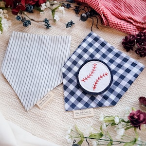 May include: Two dog bandanas. One is white with navy blue vertical stripes. The other is navy blue and white gingham with a baseball patch. Both have a small tag that reads "Olive And Cooper Co."