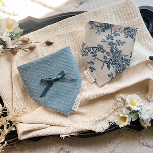 May include: Two dog bandanas, one blue with white polka dots and a blue ribbon, and one beige with a floral pattern in blue. Both bandanas have a small tag with the text "The Bark & Co."
