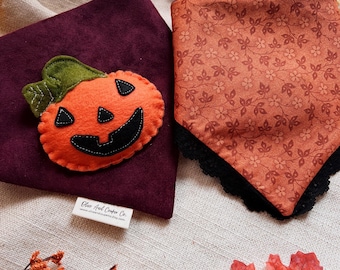May include: Two Halloween-themed pet bandanas. One is a deep purple with an orange felt pumpkin applique. The other is orange with a floral pattern and black lace trim. Fall leaves and sprigs surround the bandanas.