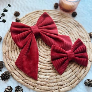 May include: Two red velvet bows, one large and one small, are displayed on a woven straw mat. The bows are made of a soft, textured fabric.