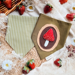 May include: Two dog bandanas. One is olive green with a white stripe pattern. The other is olive green with a felt mushroom applique. Strawberries and daisies are scattered around the bandanas.