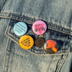 Könnte beinhalten: Nahaufnahme einer Jeansjackentasche mit fünf farbenfrohen Ansteckbuttons. Die runden Buttons zeigen Texte wie "KEEP IT QUEER" und "BUY ART MADE BY SOMEONE NOT SOME THING". Die Buttons sind in verschiedenen Farben gehalten.