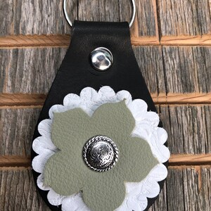 May include: Black leather keychain with a silver key ring and snap. The keychain features a white scalloped edge, a sage green leather flower, and a silver button with a floral design in the center. The background is a weathered wood surface.