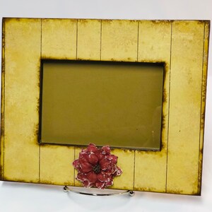 May include: A yellow wood picture frame with a red flower embellishment. The frame has a distressed look and is ready to display a favorite photo.