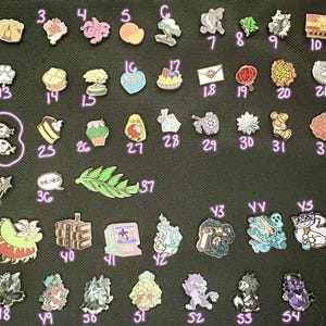 May include: A collection of assorted enamel pins arranged in rows, each labeled with a number or letter. The pins feature diverse designs including animals, plants, food items, mythical creatures, and objects, rendered in various colors and shapes.