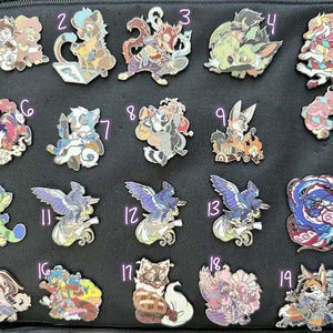 May include: A collection of nineteen colorful enamel pins featuring various fantasy creatures and characters. The pins are in a variety of shapes and sizes, with detailed designs and vibrant colors. The pins are displayed on a black surface.