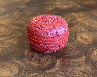 May include: A red, round, carved, wooden box with a floral design. The box is closed and has a hinged lid.