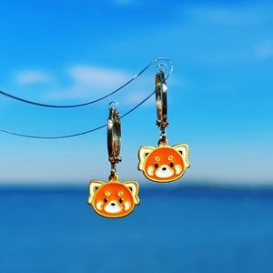 Puede incluir: Un par de pendientes de aro dorados, cada uno con un dije de panda rojo con detalles de esmalte naranja y blanco. Los pendientes cuelgan sobre un fondo de cielo azul brillante y océano.