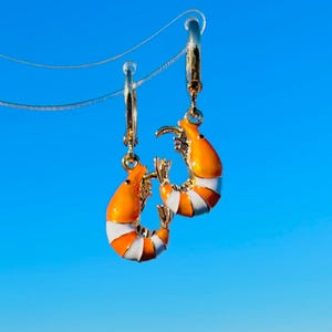Può includere: Un paio di orecchini a cerchio color oro, ciascuno con un design a gamberetto. I gamberetti sono arancioni con strisce bianche e arancioni, su uno sfondo di cielo blu brillante. Un accessorio divertente a tema nautico.
