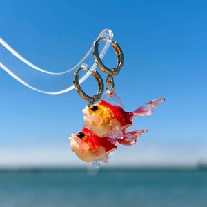 Può includere: Orecchini a cerchio dorati con un ciondolo a forma di pesce rosso rosso e giallo appeso a ogni cerchio. Gli orecchini sono fotografati su uno sfondo di cielo blu e oceano.