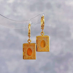 Könnte beinhalten: Goldene Creolen mit einem kleinen goldenen Anhänger in Form eines Medaillons, der an jeder Creole hängt. Die Medaillons haben ein detailliertes Design und eine ovale Öffnung.